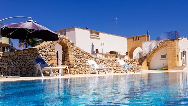 Une piscine à l’eau d’un bleu cristallin, entourée de chaises longues blanches et d’un mur de pierre.