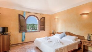 Une chambre à coucher avec un lit, une télévision, une fenêtre donnant sur des palmiers et un sol carrelé.