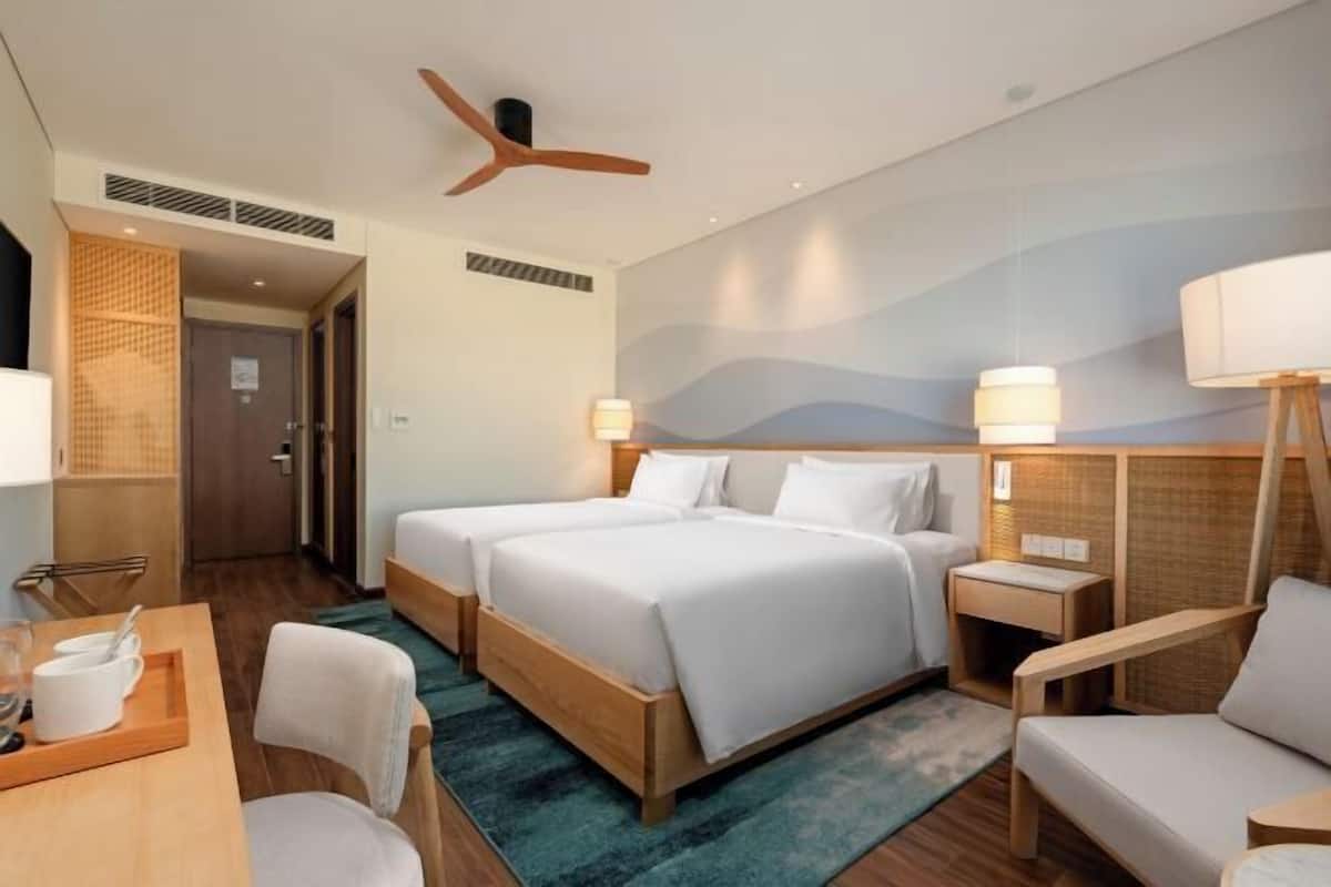 A hotel room with a large bed, two bedside tables, a desk with a chair, a standing lamp, and a ceiling fan.