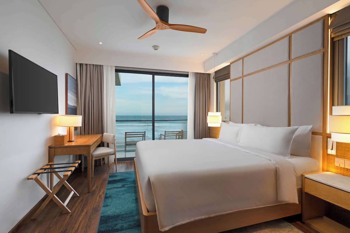 A hotel room with a large bed, a desk with a chair, a TV, and a view of the ocean.