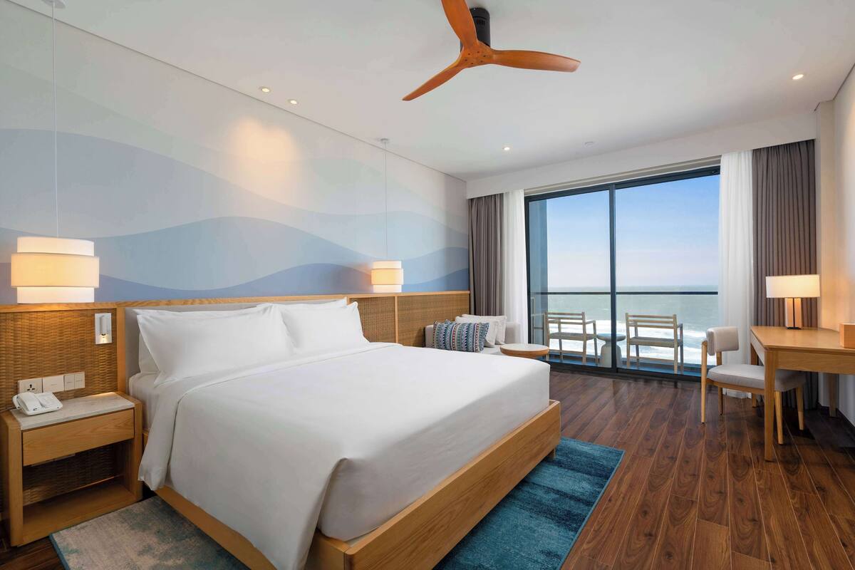 A modern hotel room with a large bed, wooden headboard, a desk, and a view of the ocean through large windows.