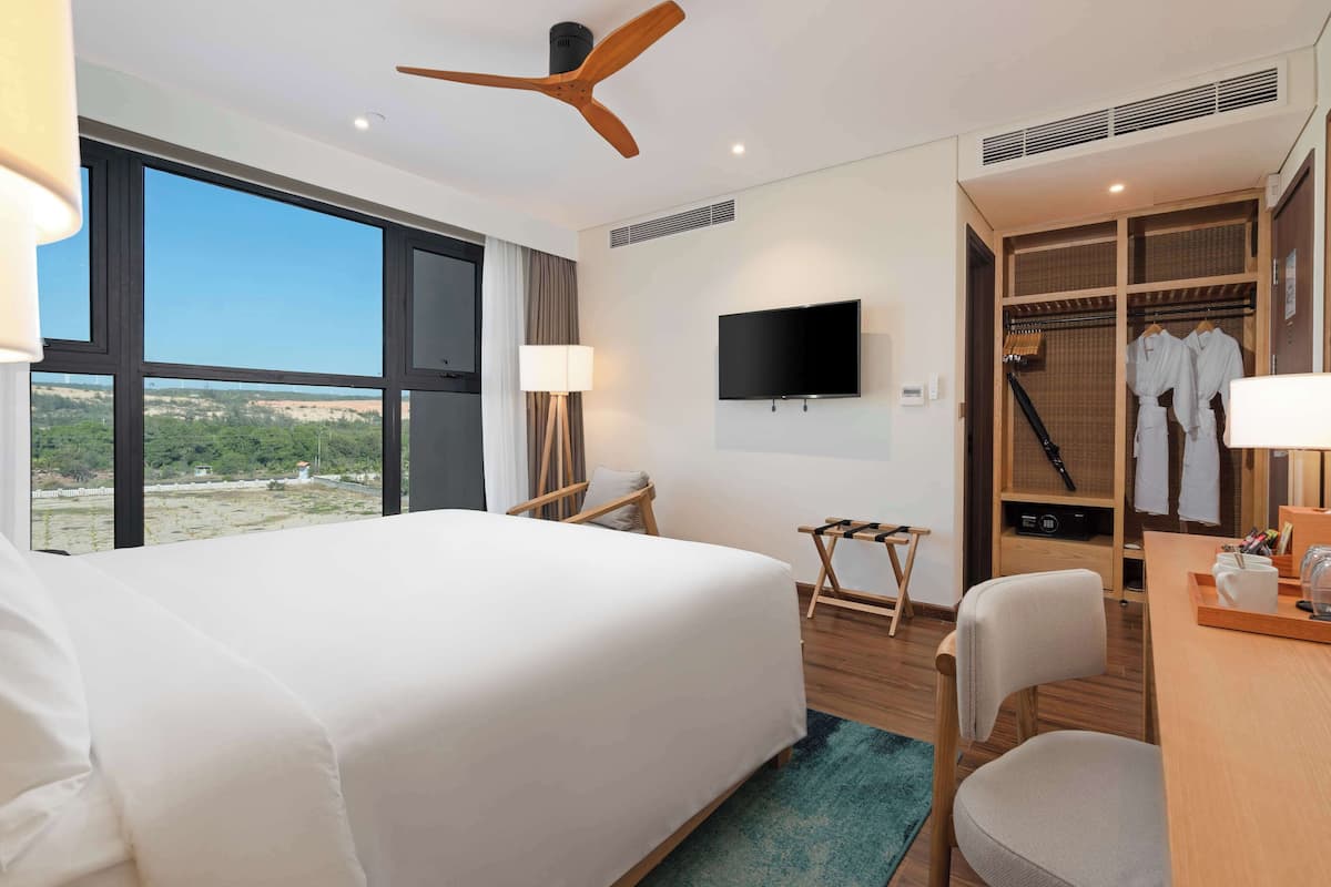 A hotel room with a large bed, a desk, a TV, a wardrobe, and a view of the landscape.