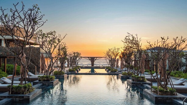 A pool area with lounge chairs and umbrellas, a view of the sunset, and a building in the background.
