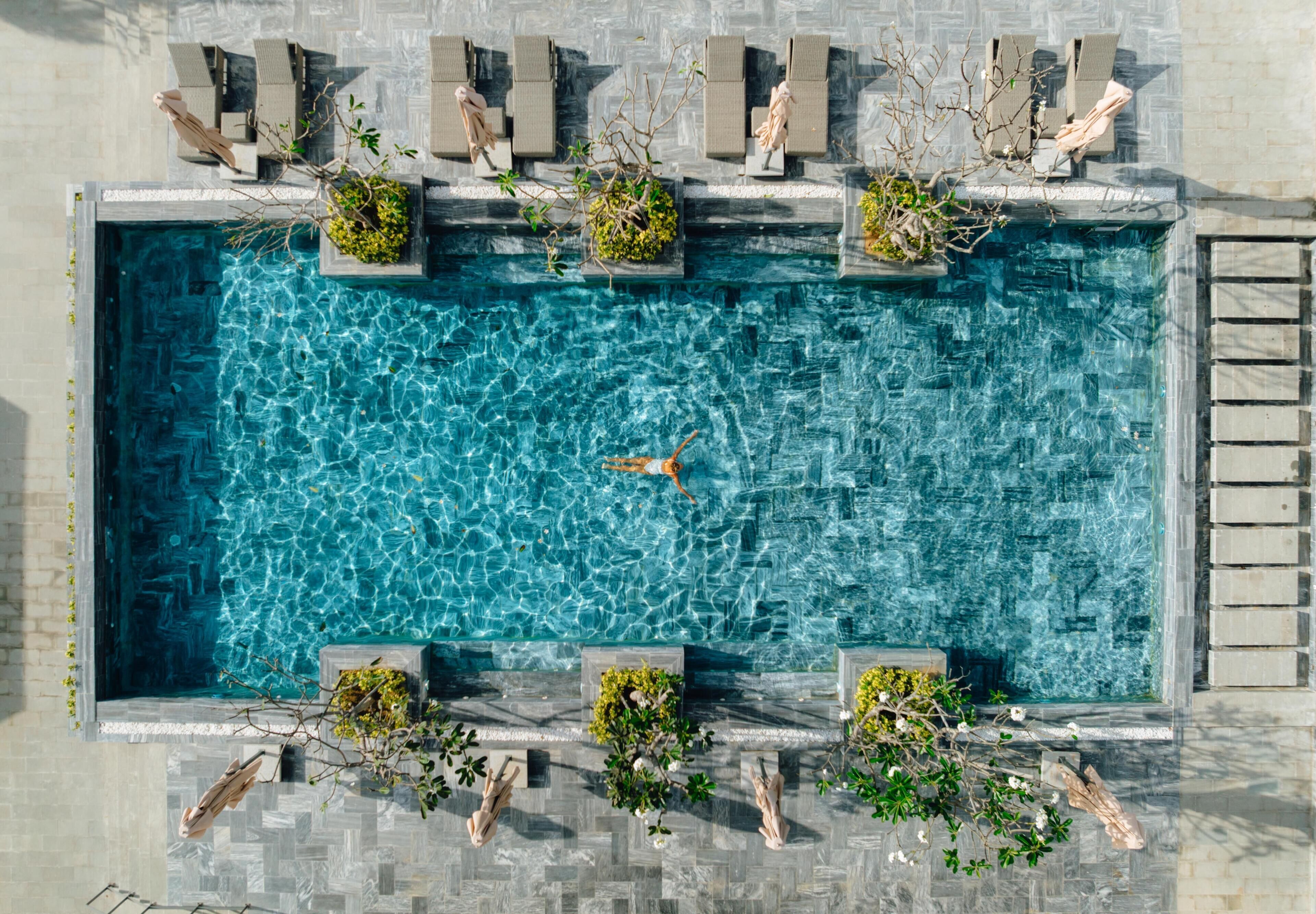 A rectangular swimming pool with clear blue water, surrounded by stone tiles and equipped with multiple lounge chairs and planters with greenery.