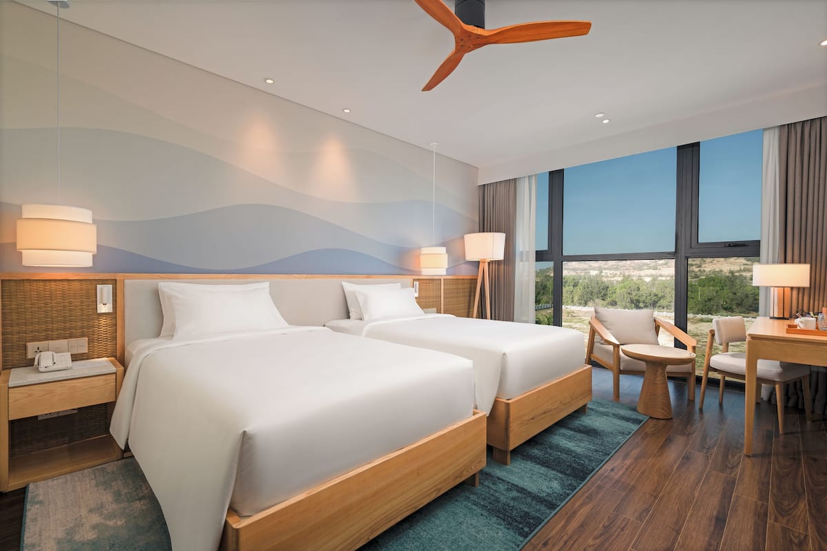 A hotel room with two beds, a desk, a chair, and a view of the outdoors.