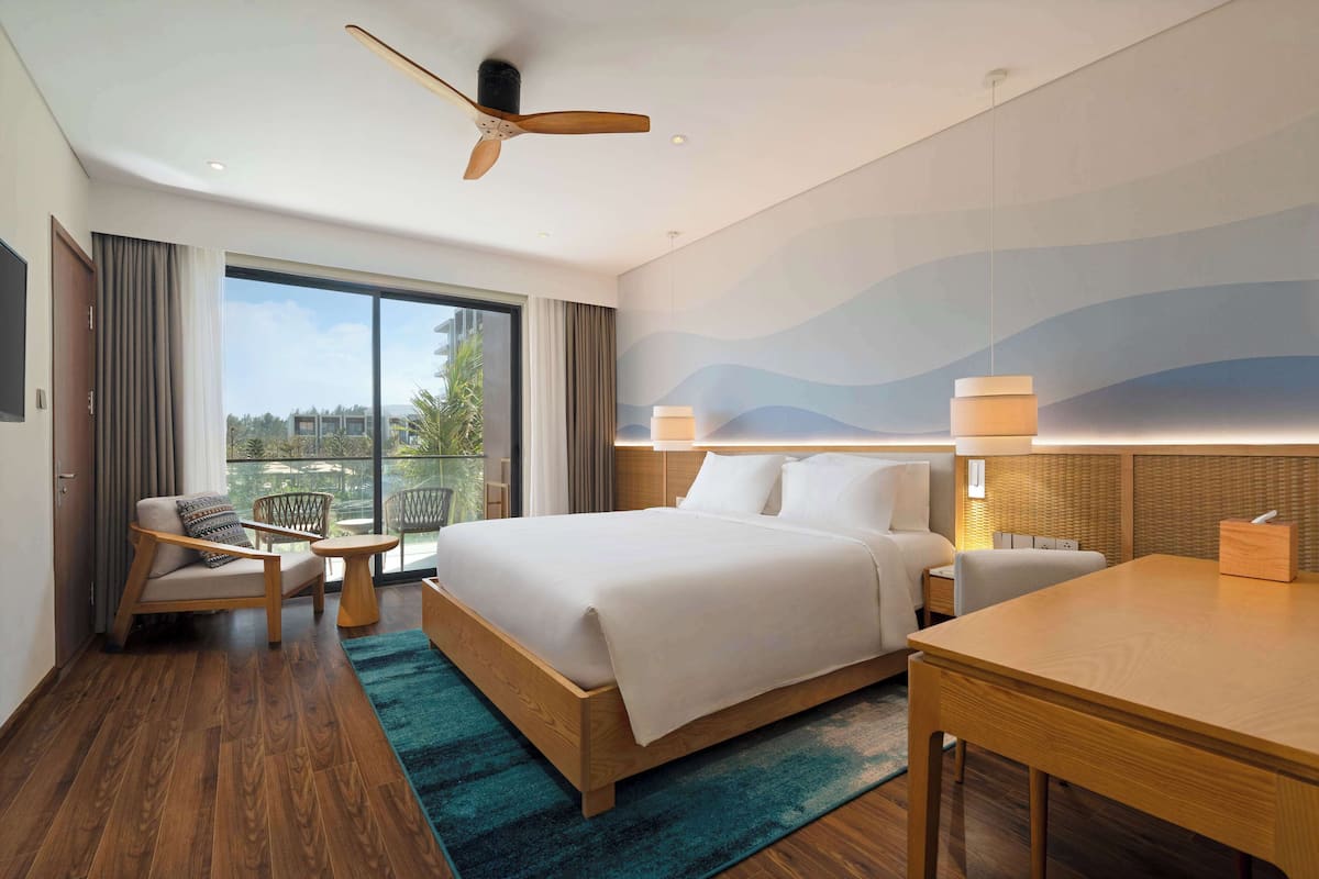 A modern hotel room with a large bed, a desk, a chair, a small table, and a view of the outdoors.