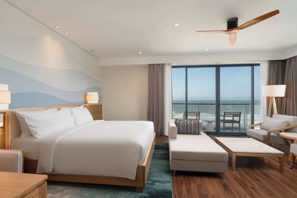 A modern hotel room with a large bed, a sitting area with a sofa and armchairs, a ceiling fan, and a view of the ocean through large windows.