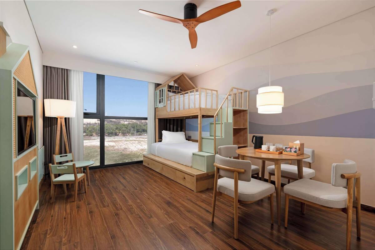 A hotel room with a wooden floor, a bunk bed, a desk with chairs, a lamp, and a view of the outdoors.