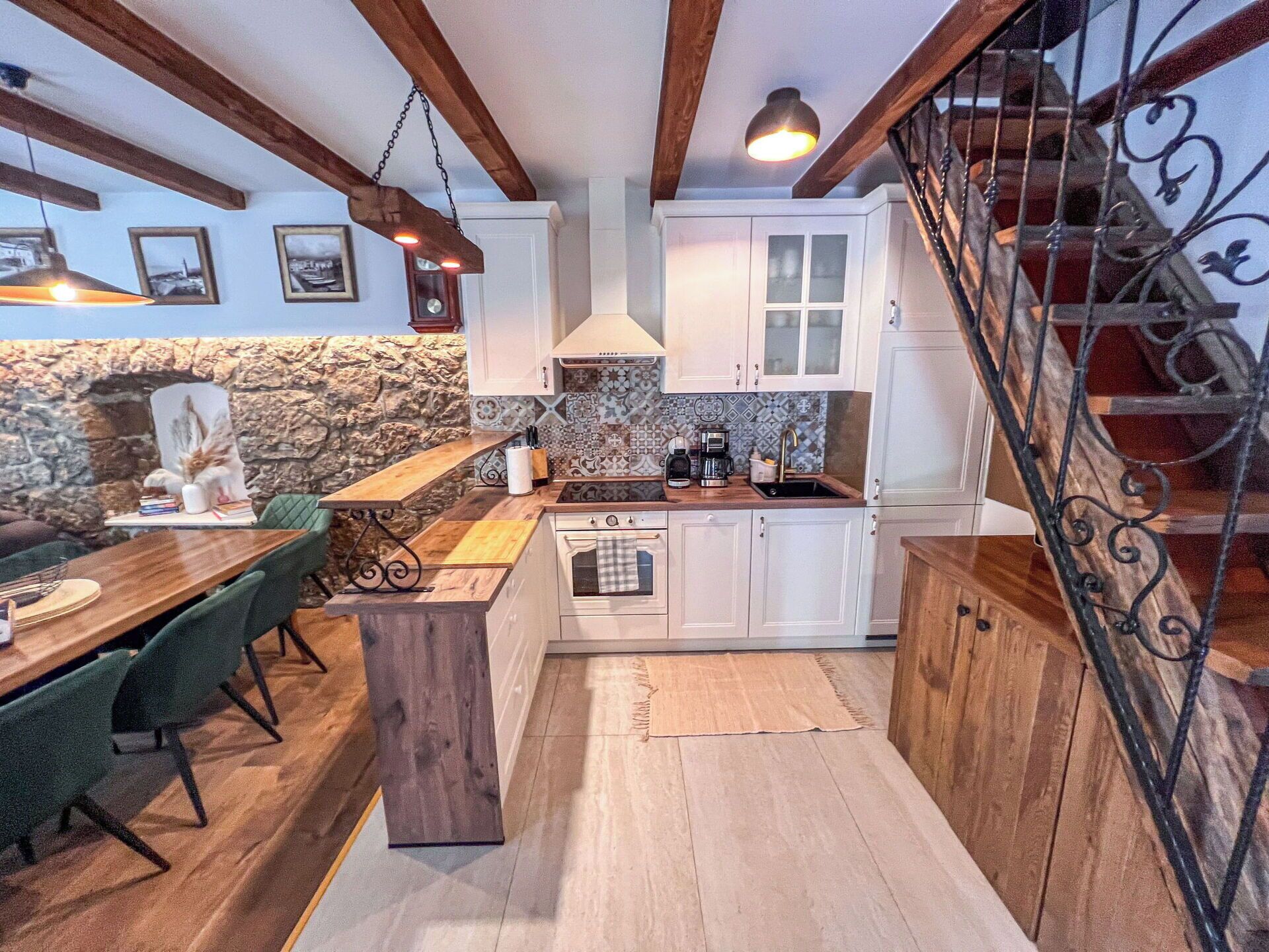 Una cocina con vigas de madera, armarios blancos, pared de piedra, zona de comedor con sillas verdes y una escalera con barandilla de hierro forjado.