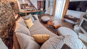 Ein Wohnzimmer mit einer Couch, einem Hocker und einem kleinen Holztisch. Im Hintergrund ist eine Küche mit Essbereich zu sehen.
