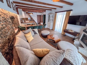 A living room with a sofa, ottoman, and a small wooden table. There is a kitchen in the background with a dining area.