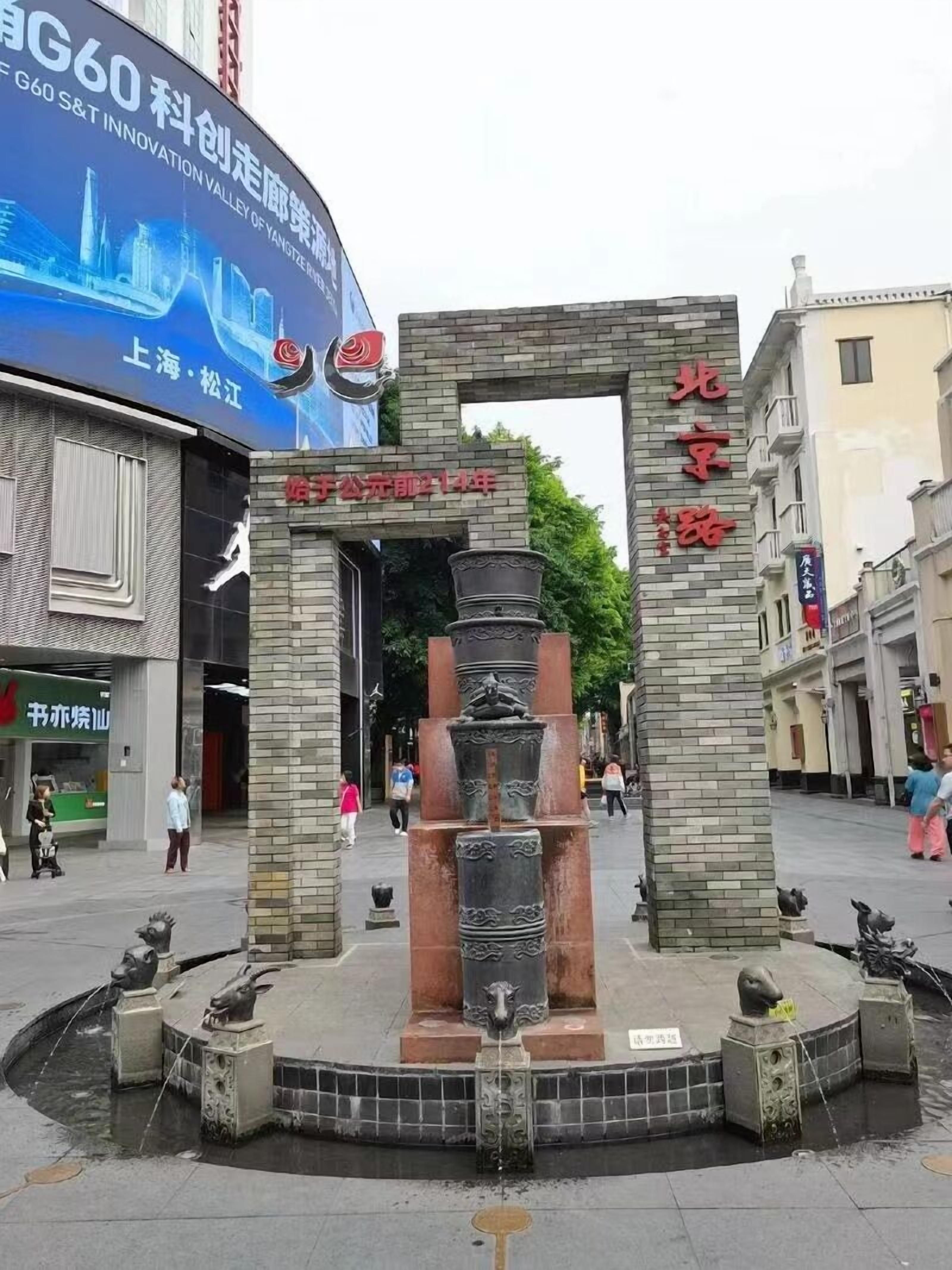 A public square with a central fountain, surrounded by stone pillars with inscriptions. There is a large billboard in the background advertising a company.