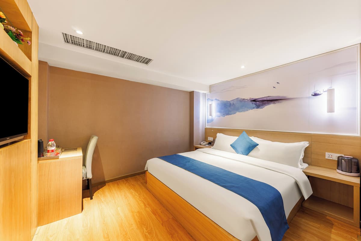 Una habitación de hotel con cama, escritorio, silla, televisor de pantalla plana, mesita de noche y una obra de arte en la pared.