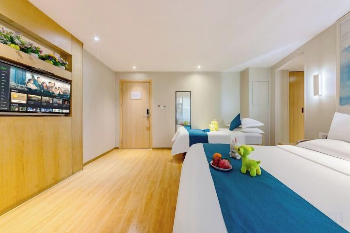 Una camera d'hotel con pavimento in legno, un letto con copriletto blu, un secondo letto, una scrivania con una sedia e una televisione a parete.