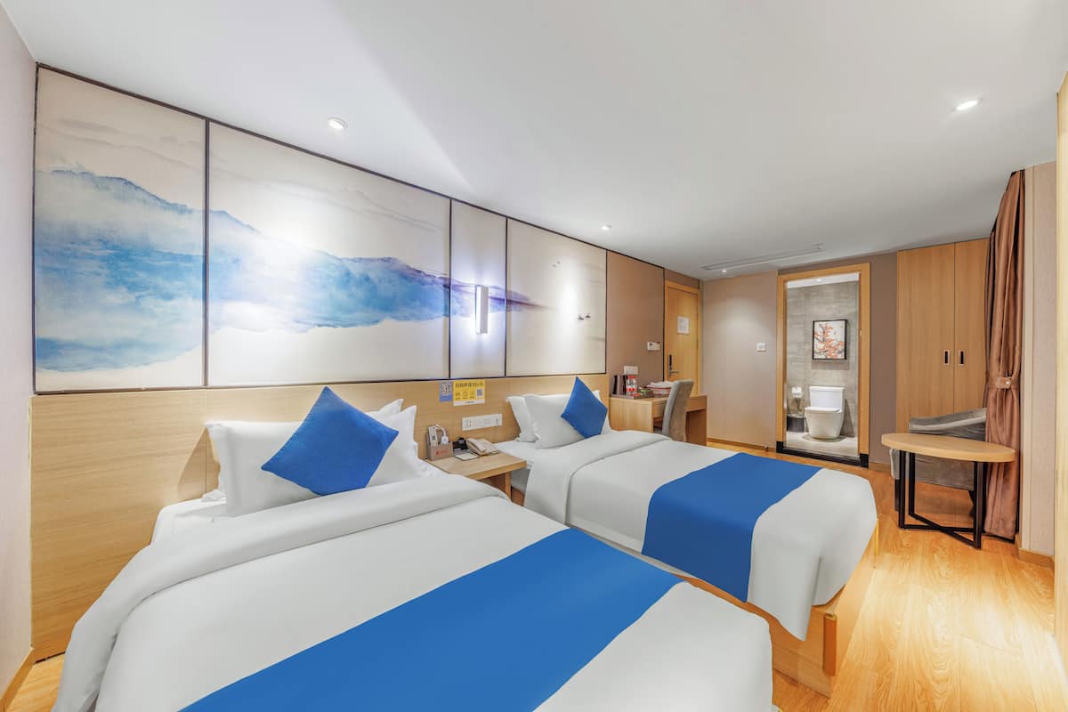 Habitación de hotel con dos camas, cabecero de madera, un cuadro abstracto grande, una mesita y un baño visible a través de una puerta abierta.