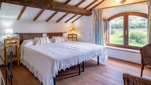 Une chambre avec un plafond en bois, un lit recouvert de linge de lit blanc, une table de chevet, une lampe et une vue sur la verdure par la fenêtre.