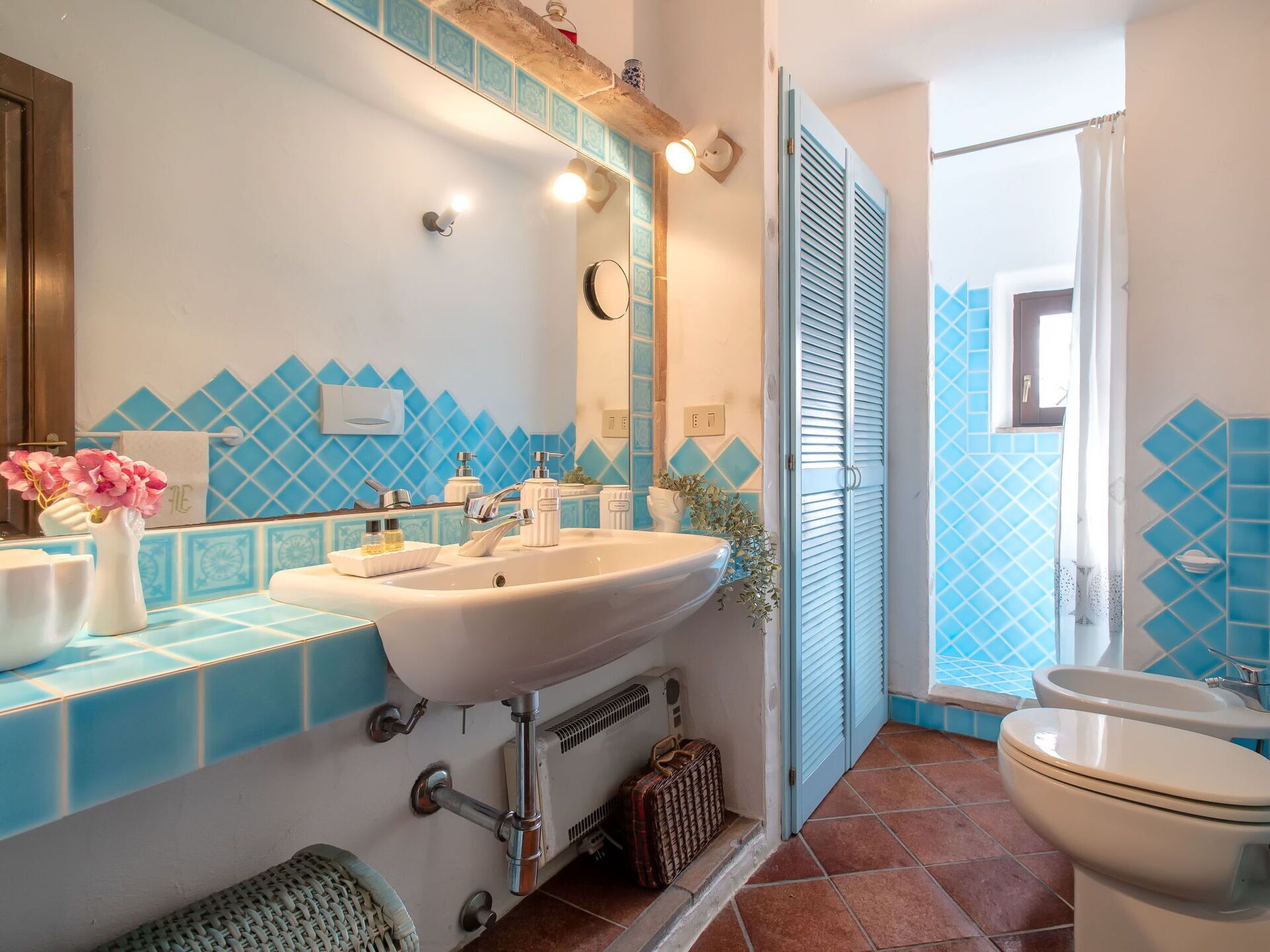 Ein Badezimmer mit einem weißen Waschbecken, zwei Wasserhähnen, einem Spiegel und einer Toilette. Ein blauer Fliesenverkleidung und ein Fenster mit Vorhang sind ebenfalls vorhanden.