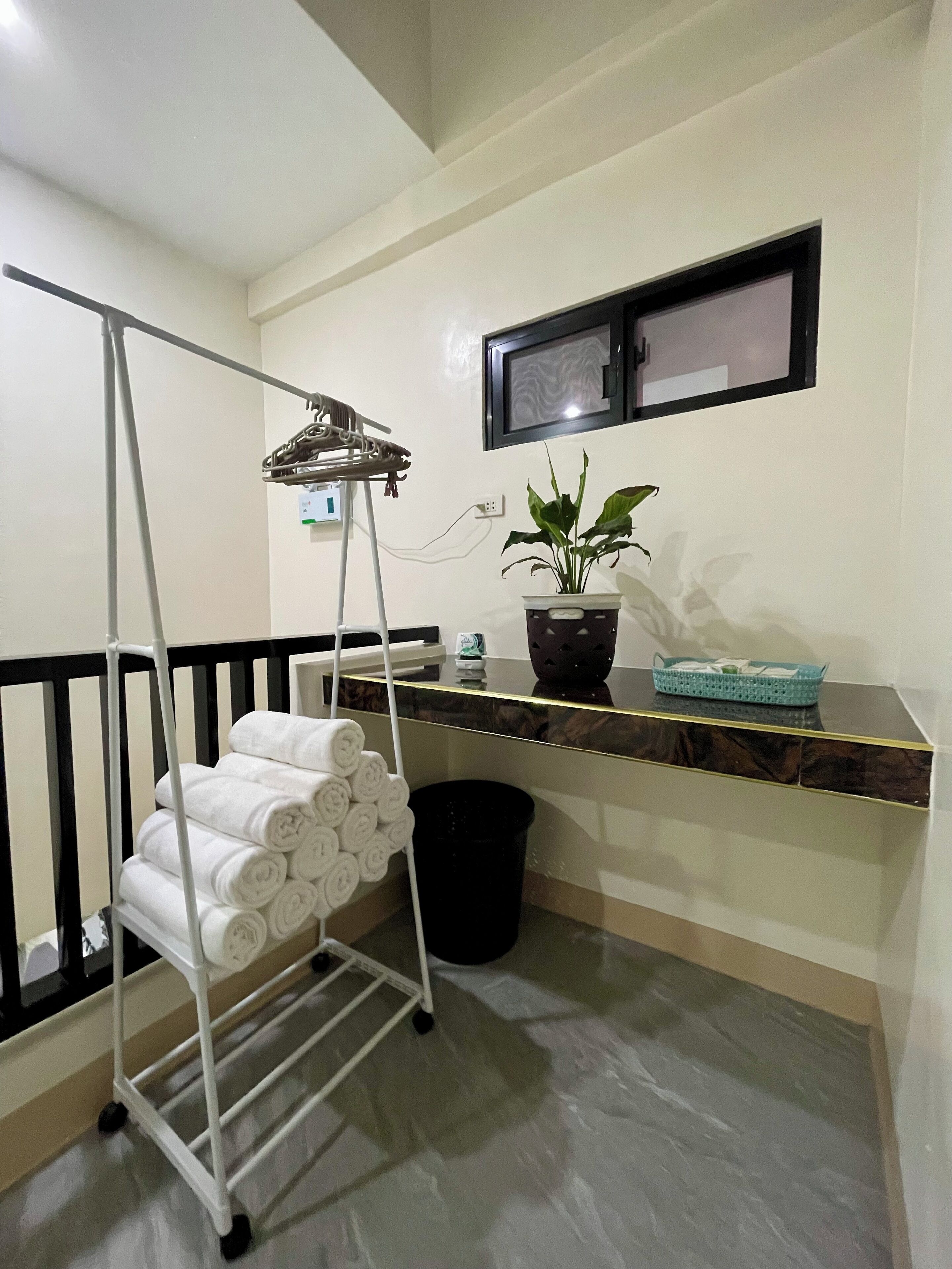 A room with a white metal drying rack holding rolled towels, a potted plant on a shelf, and a black trash bin.