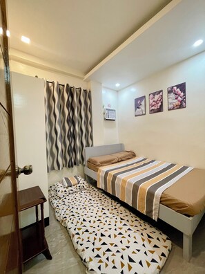 A bedroom with two beds, a bedside table, a wall-mounted air conditioner, and three framed pictures.