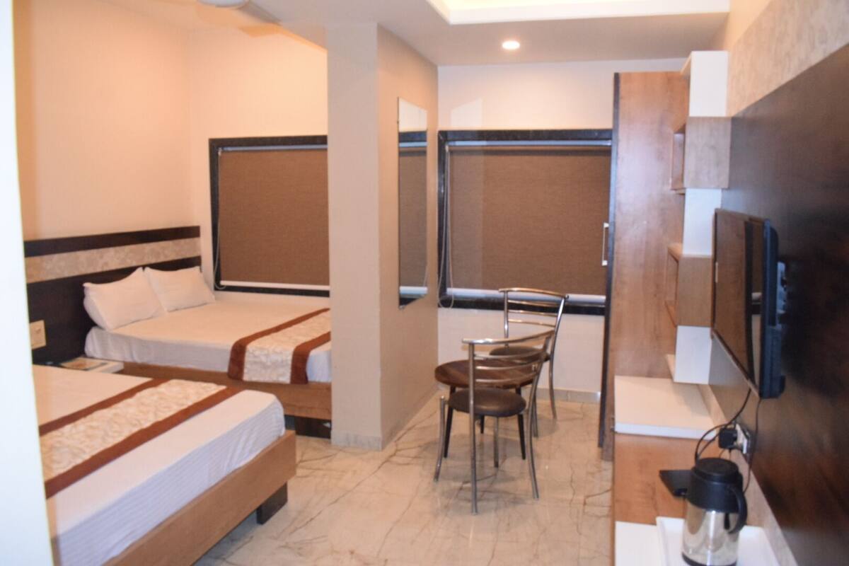 A hotel room with two single beds, a small table with a chair, a TV mounted on the wall, and a window with a view.