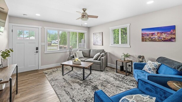 A living room with a grey sofa, blue sofa, and a coffee table. There are two windows with blinds, a ceiling fan, and a painting on the wall. - Renovated Urban Oasis in Indy (Indianapolis)