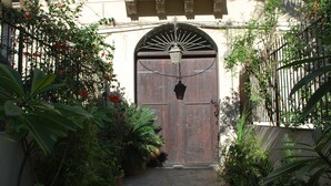 A narrow pathway leading to a wooden door with a metal arch above it, surrounded by lush greenery and a hanging light fixture. - Volta House (Palermo)