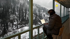 A person sitting on a chair by a window with a laptop, overlooking a snowy landscape with trees and a cloudy sky.