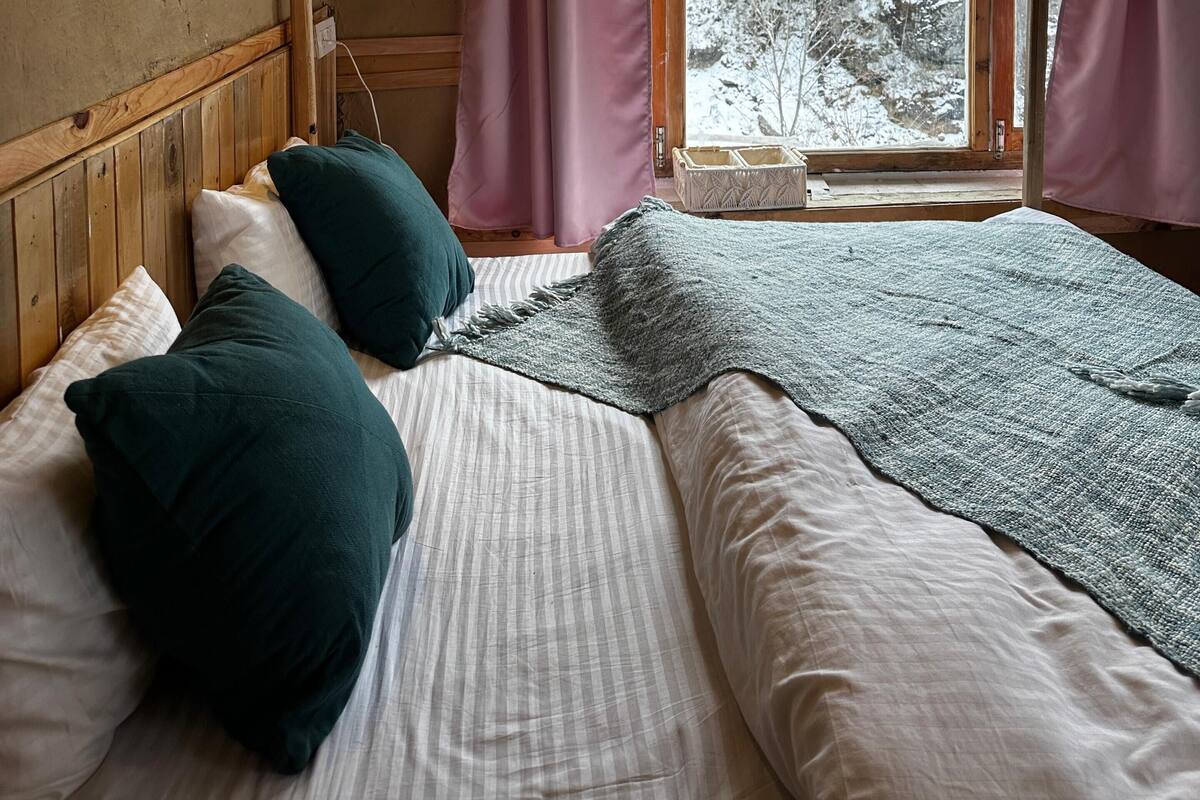 A cozy cabin room with a bed, wooden walls, a window with curtains, and a view of a snowy forest.