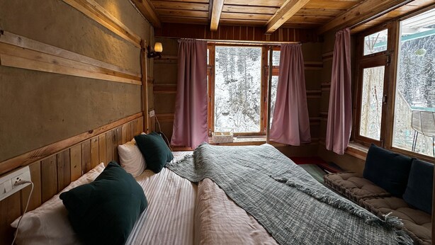 A wooden cabin room with a bed, a window with curtains, and a view of a snowy forest.