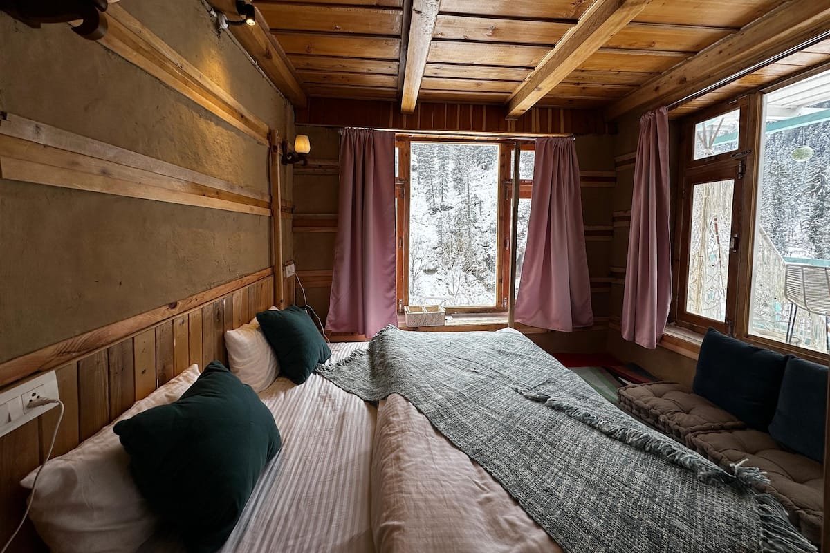 A wooden cabin room with a bed, a window with curtains, and a view of a snowy forest.