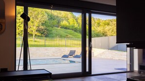 Une chambre moderne dotée d’une grande porte-fenêtre coulissante qui donne sur un jardin avec une piscine, des chaises longues et une vue sur un paysage verdoyant.
