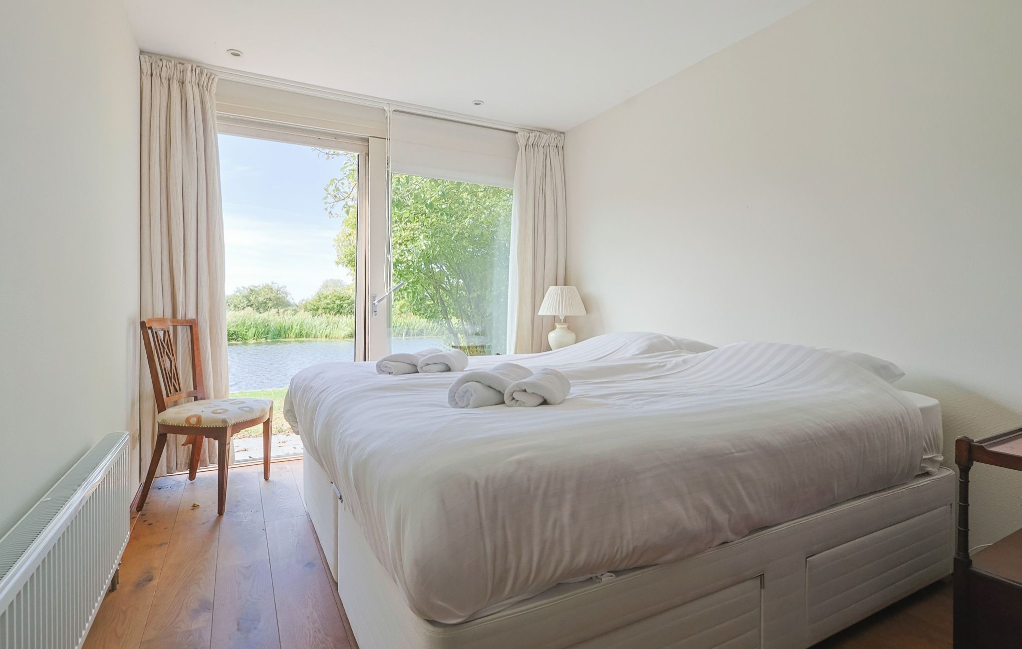 Ein Schlafzimmer mit einem großen Bett, einem Sessel, einer Lampe und Blick auf Grün durch das Fenster.