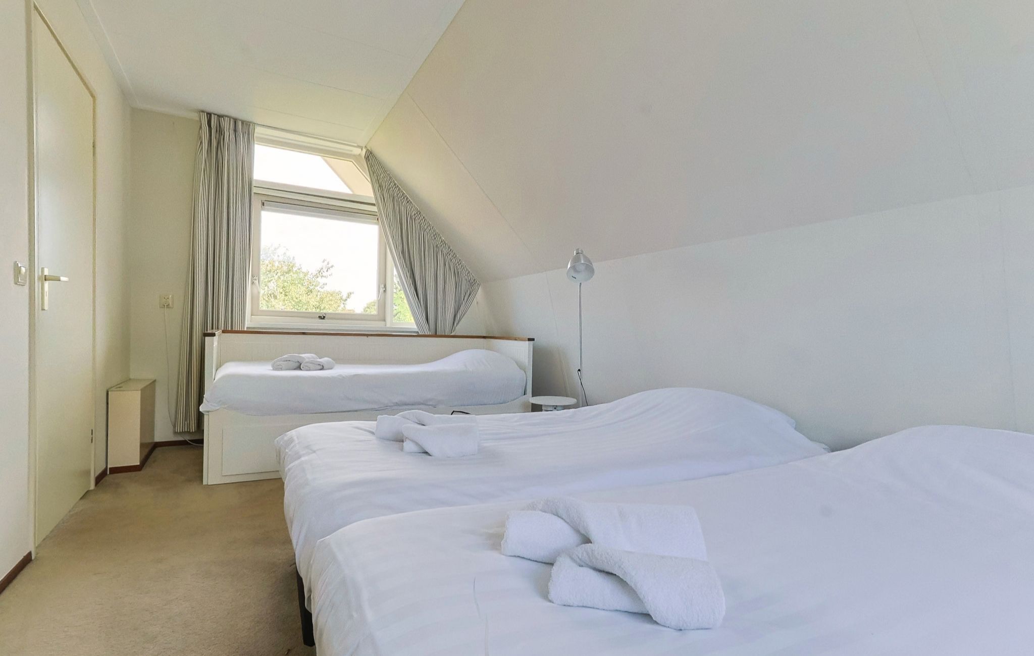 Ein Hotelzimmer mit zwei Betten, einem Fenster mit Vorhängen, einer Nachttischlampe und einem kleinen Tisch.
