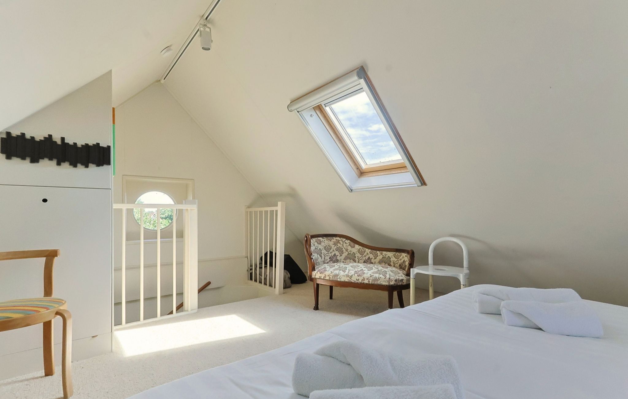 A bedroom with a sloped ceiling, a skylight, a bed with white bedding, a wooden chair, and a white bedside table.