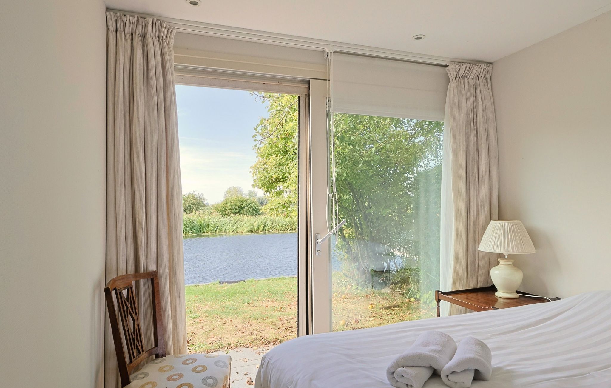 Ein Schlafzimmer mit einem Bett, einem Sessel, einem Nachttisch mit Lampe und Seeblick durch eine Glastür.