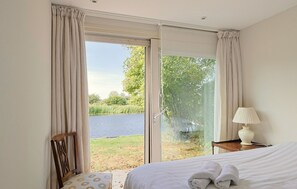 A bedroom with a bed, a chair, a bedside table with a lamp, and a view of a lake through a glass door.