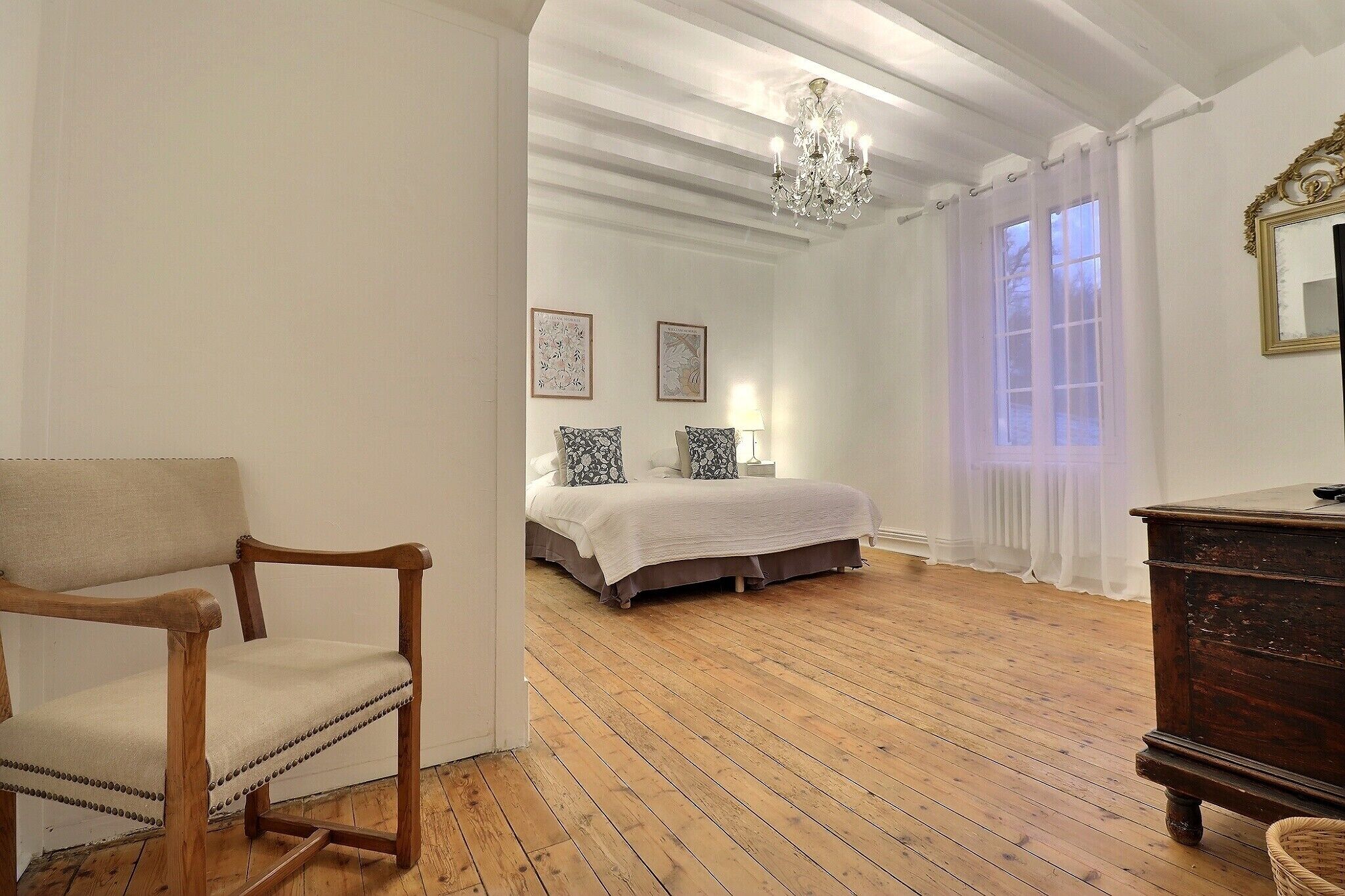 Una stanza con pavimento in legno, un letto con copriletto bianco, una sedia di legno e un comò.