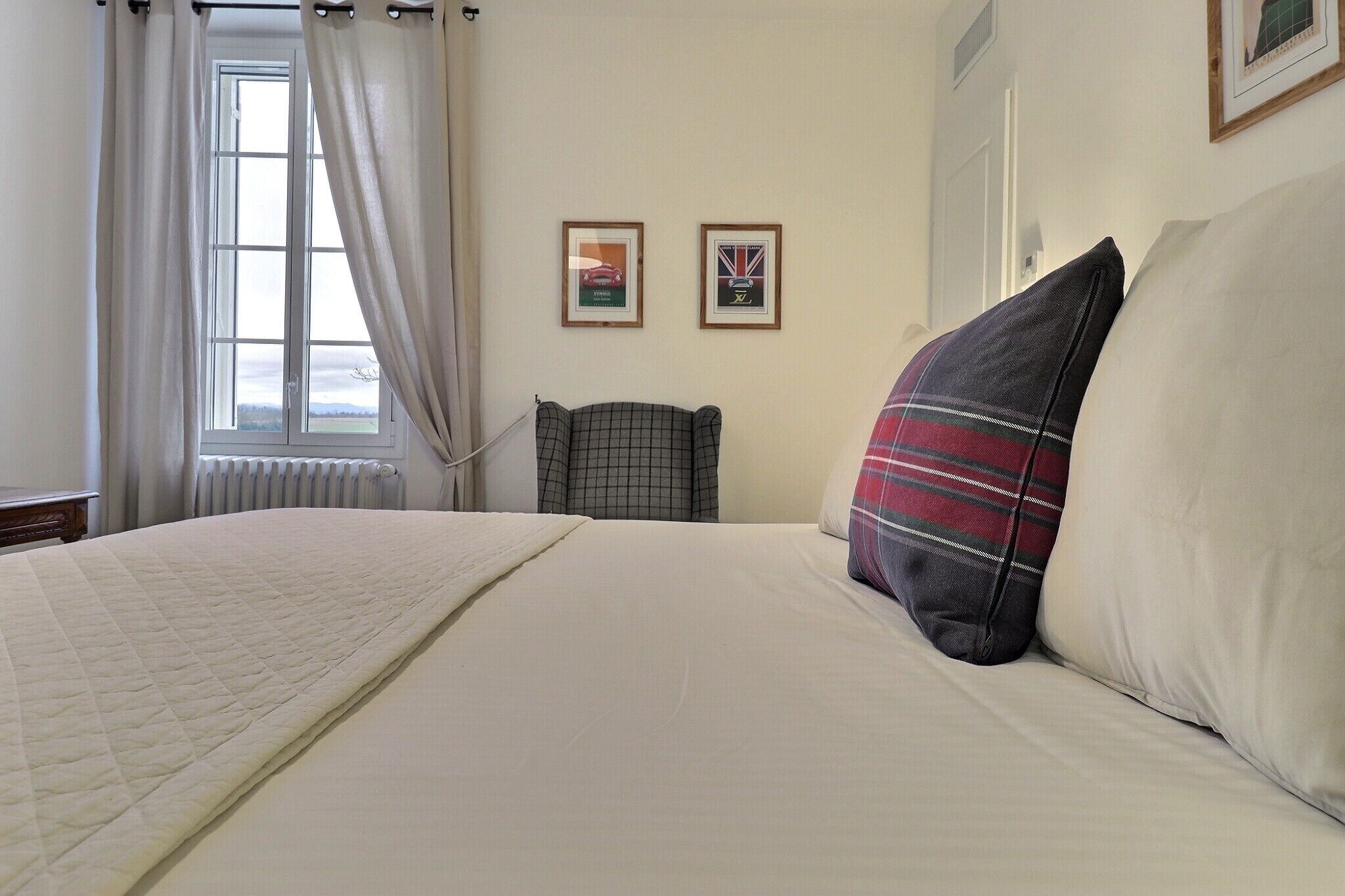 A neatly made bed with a quilted blanket, a plaid pillow, and a plain pillow. There is a window with curtains, a radiator, and two framed pictures on the wall.