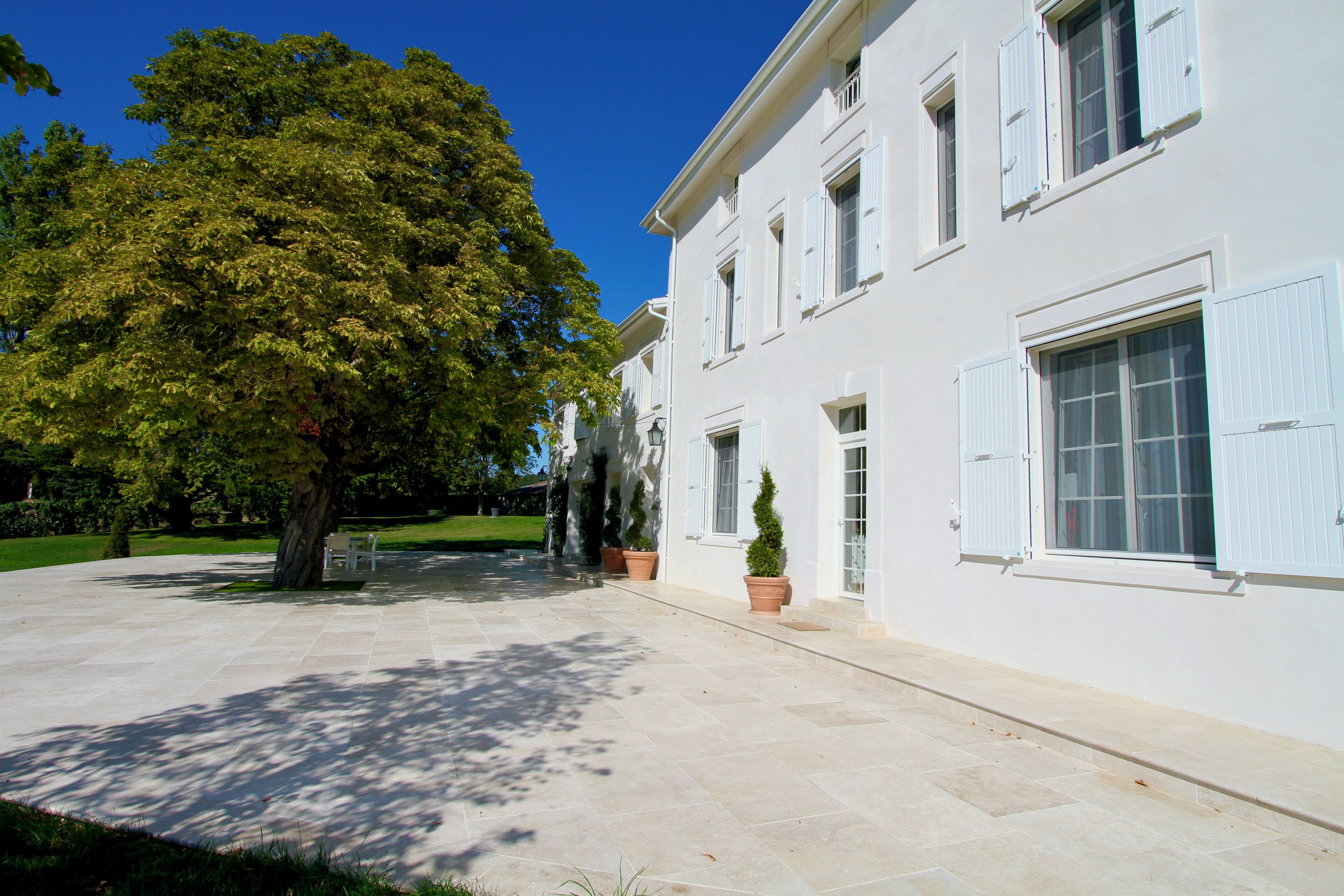 Un edificio bianco con numerose finestre e persiane, un cortile pavimentato e un grande albero.