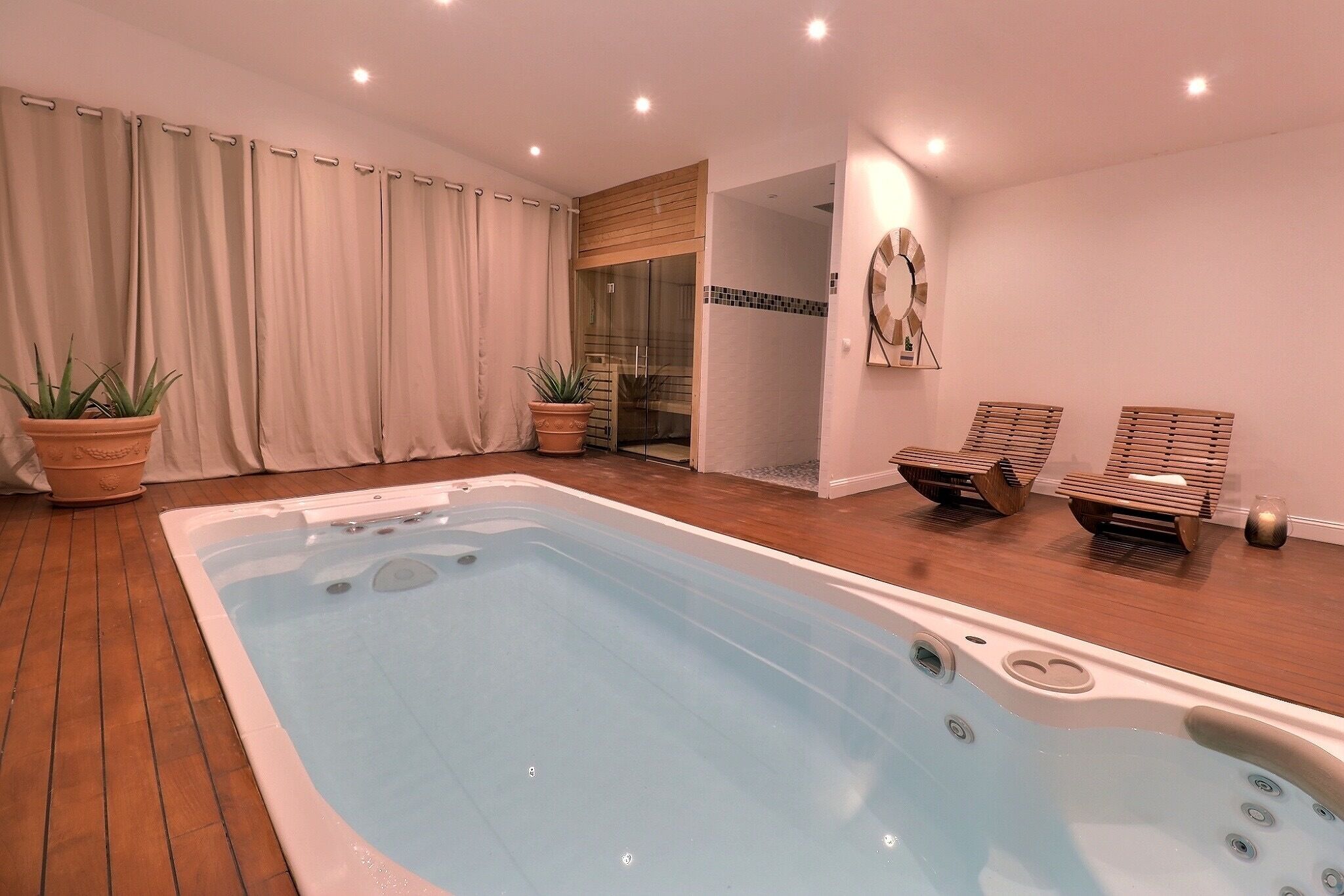 Un espace piscine intérieur moderne, doté d’un jacuzzi, d’un plancher en bois, de plantes en pot et d’une porte en verre donnant accès à un espace douche.