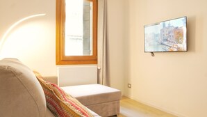 A room with a sofa, a small ottoman, a window with curtains, and a wall-mounted TV displaying a cityscape. - The Glass House (The Art of Murano) Apartment in the heart of Venice (Venezia)