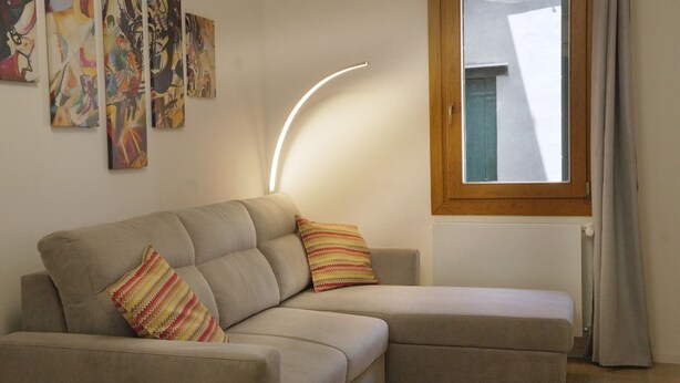 A modern living room with a beige sofa, two patterned throw pillows, abstract wall art, a curved light fixture, and a window with a view of a building. - The Glass House (The Art of Murano) Apartment in the heart of Venice (Venezia)