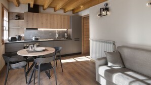 Een moderne keuken met houten balken, een eethoek met een ronde tafel en stoelen, en een grijze bank in een lichte kamer met natuurlijk licht.