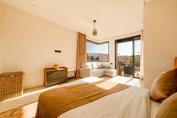 A modern hotel room with a large bed, a sofa, a small table, and a view of the city through the window.
