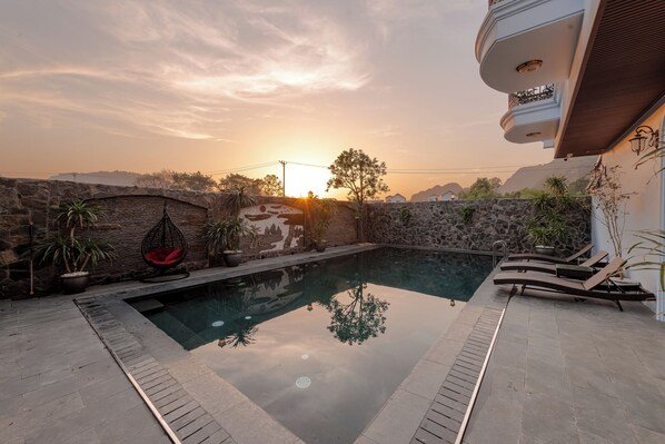 Una piscina con pared de piedra y plantas en macetas, una silla colgante roja y un balcón con barandilla.