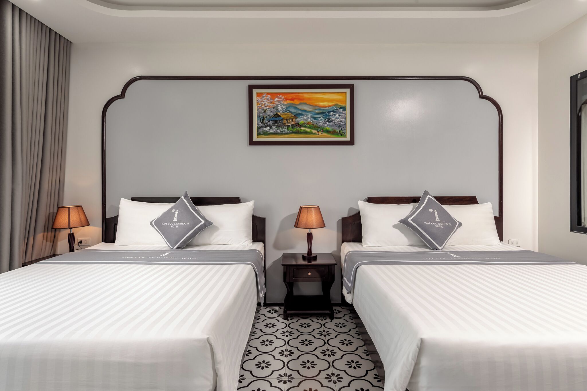 A hotel room with two beds, a nightstand with lamps, a framed painting on the wall, and patterned floor tiles.