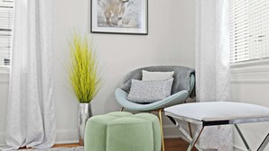 Una habitación con un sillón gris, un banquillo verde, una tabla de planchar blanca y un jarrón plateado con plantas verdes.