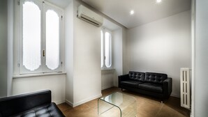A modern living room with a black leather sofa, a glass coffee table, and two windows with frosted glass panes. - Trinità Grand Suite (Rome)