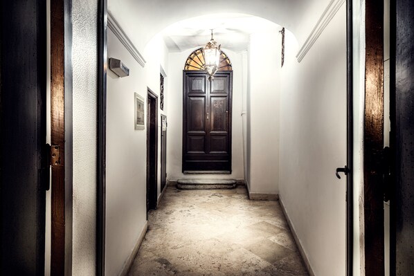 Um corredor estreito com uma porta de madeira escura no final, uma parede branca e um chão texturizado.