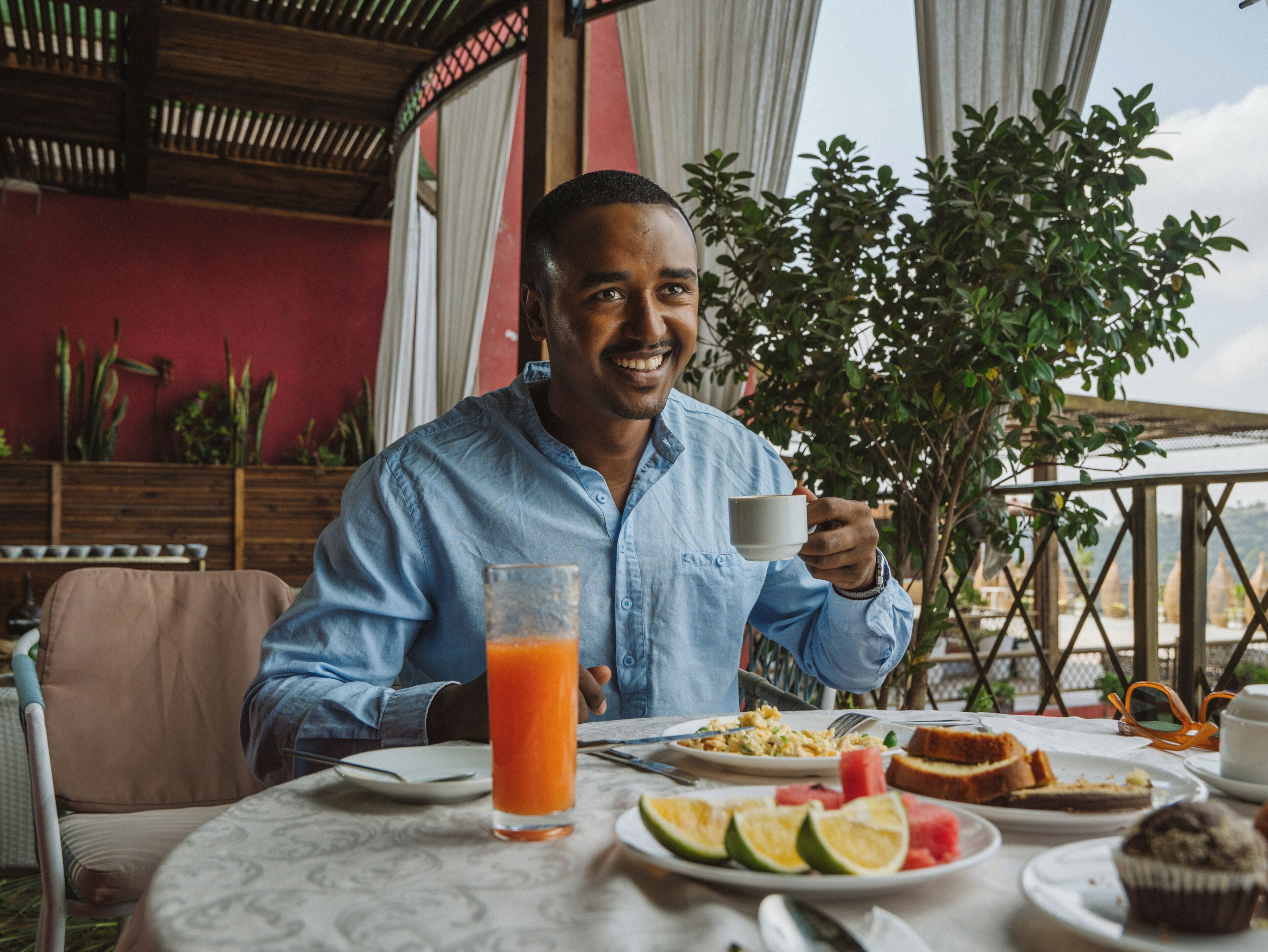 A person seated at an outdoor table with a meal, a drink, and a view of greenery and buildings.